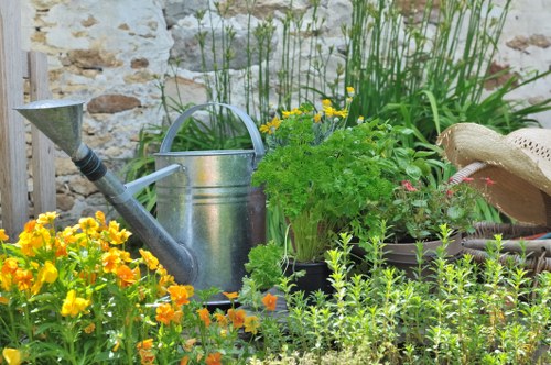 The image depicts a small, well-organized garden corner featuring a variety of plants and garden tools. In the foreground, there are vibrant yellow and orange flowers, likely marigolds, growing densely alongside lush green foliage. Behind these flowers, a collection of potted plants with different textures and shades of green is arranged, including some with pink and white blossoms. To the left, a weathered metal watering can is positioned on the ground, with its spout directed towards the plants, indicating recent or upcoming watering activity. To the right, a natural straw hat rests on the edge of a plant pot, suggesting someone is tending to the garden. The background includes a partial view of a textured stone or brick wall, typical of a traditional UK garden setting, with taller garden greenery behind the pots, such as ornamental grasses. The scene appears to be outdoors during daylight hours, with bright, natural light illuminating the plants and tools, emphasizing the healthy, thriving condition of the garden. This scene aligns with gardening and outdoor maintenance services in the Harrow area, highlighting typical domestic gardening elements, including flower beds, potted plants, and garden accessories, carefully arranged in a cozy and practical garden space.