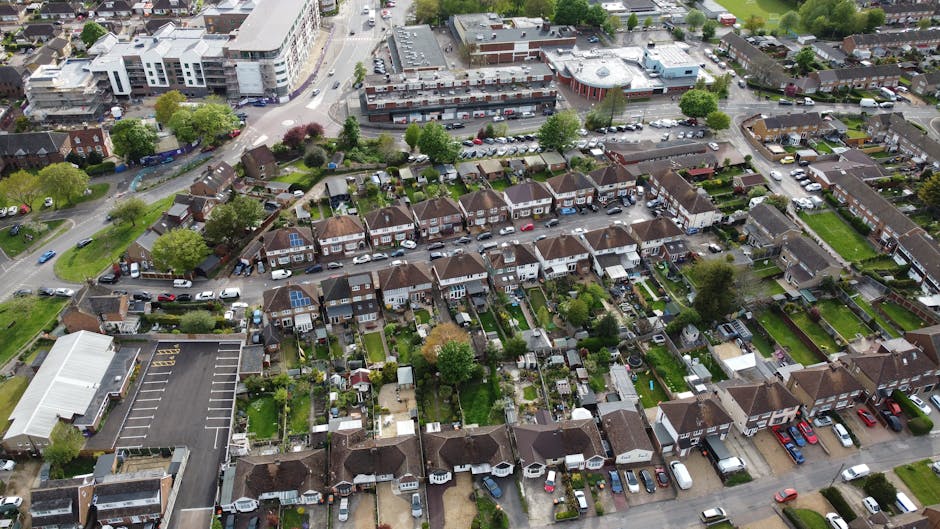 An aerial view of a residential suburb with rows of terraced houses featuring small front gardens, driveways, and well-maintained lawns. The streets are lined with parked cars, and leafy trees are sca
